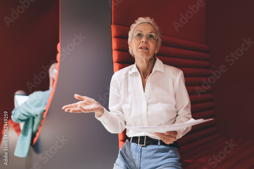 Communicative senior woman holding papers, gesturing in discussion, with a colleague's presence in a vibrant red office space