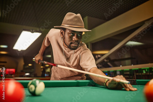 Dramatic portrait of adult African American man playing pool and hitting ball with cue stick in pool club copy space