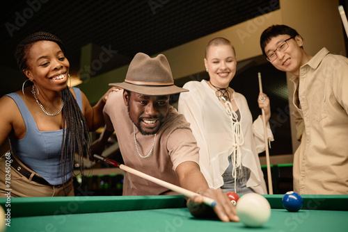 Multiethnic group of friends playing pool together with focus on smiling Black man hitting ball with cue stick copy space