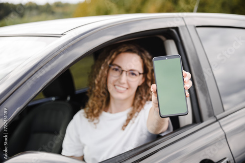 young beautiful smiling woman in car looking straight to camera, holding smartphone with green screen in hand, attractive caucasian woman in white t-shirt, selective focus
