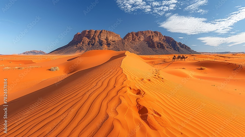   A group rides camels through a sandy desert, blue sky overhead, mountains distant backdrop
