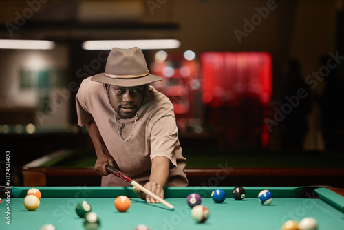 Waist up portrait of adult African American man wearing hat and hitting ball with cue stick while playing pool copy space