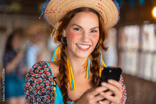 Delighted young lady in traditional Brazilian costume interacting with her smartphone at a Junina celebration.