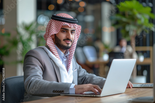 Smiling Businessman in Traditional Arab Attire Working on Laptop in Modern Office photo