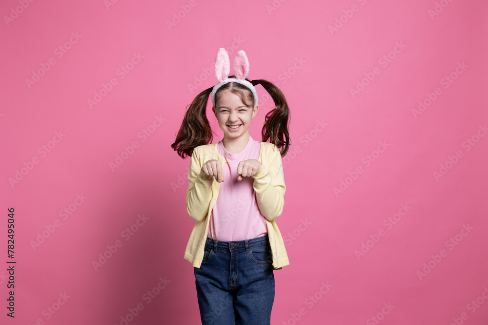 Adorable little girl spending Easter holiday by hopping around like a rabbit for the camera while wearing bunny ears. Delighted happy youngster playing and chuckling on pink background.
