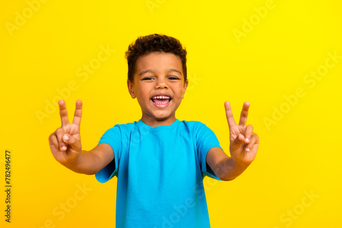 Photo of friendly pleasant cute child with curly hair dressed blue t-shirt showing v-sign symbol isolated on vibrant yellow background