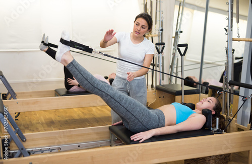 Young woman practicing Pilates system on reformer supervised by qualified trainer.