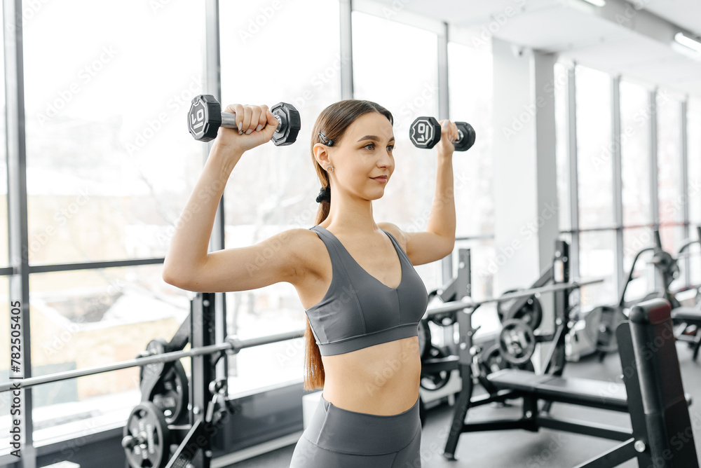 young athletic caucasian woman trains in fitness gym, standing and doing dumbbell press up, brunette girl in grey top and leggings, healthy lifestyle concept young athletic caucasian woman trains in fitness gym, standing and doing dumbbell press up, brunette girl in grey top and leggings, healthy lifestyle concept