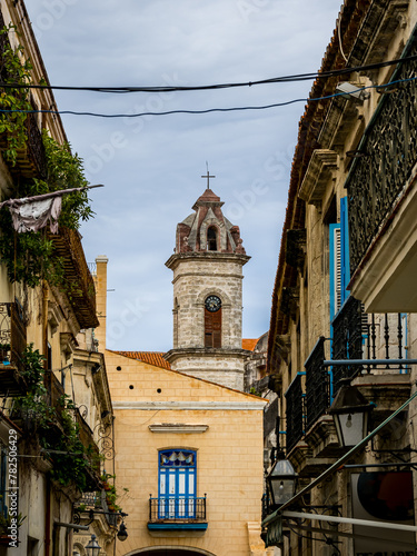 Portrait view of Havana Cathedral baroque bell tower, framed by the ornate balconies of Calle de San Ignacio street, offers a glimpse into the colonial charm of Old Havana, Cuba, on an overcast day.