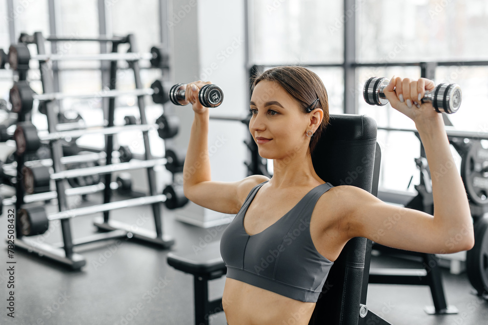 young athletic caucasian woman trains in fitness gym, doing dumbbell press up on bench, brunette girl in grey top and leggings, healthy lifestyle concept young athletic caucasian woman trains in fitness gym, doing dumbbell press up on bench, brunette girl in grey top and leggings, healthy lifestyle concept