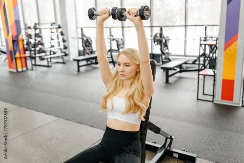 young athletic caucasian woman trains in fitness gym, dumbbell press up on bench, blond girl in white top and black leggings, healthy lifestyle concept