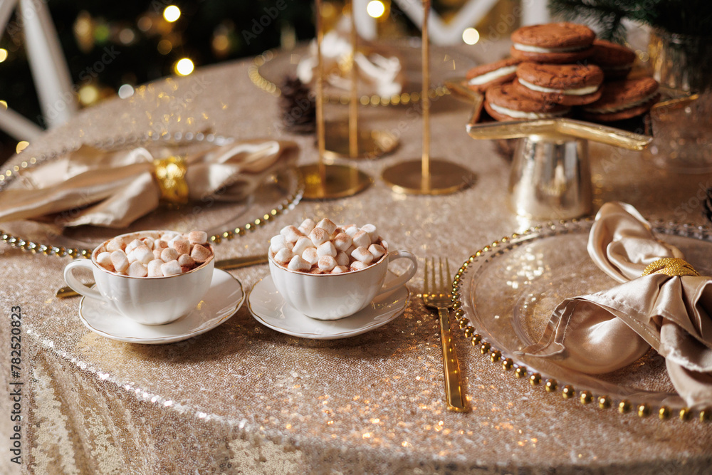 Christmas or New Years festive party luxury table setting. Glittering tablecloth, cups with cacao and marshmallow, chocolate cookies, glasses plates, napkins, golden cutlery, candlestick, garlands Christmas or New Years festive party luxury table setting. Glittering tablecloth, cups with cacao and marshmallow, chocolate cookies, glasses plates, napkins, golden cutlery, candlestick, garlands