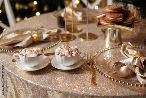 Christmas or New Years festive party luxury table setting. Glittering tablecloth  cups with cacao and marshmallow  chocolate cookies  glasses plates  napkins  golden cutlery  candlestick  garlands