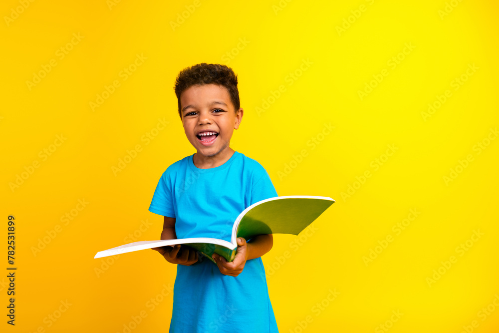 Photo of cheerful small child with curly hair dressed blue stylish t-shirt holding book having fun isolated on vibrant yellow background - obrazy, fototapety, plakaty 