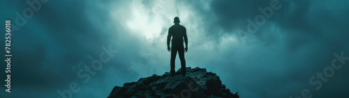 Silhouette of a man standing atop a rugged peak against a dramatic and tumultuous sky. photo