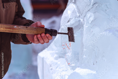 A sculptor processes a large ice cube with a sharp hand tool. Ice dust and ice fragments in the air. Ice sculpture festival in the city. Concept of hobby and interaction between man and nature