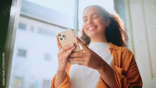 Using gadgets. Chatting online. Happy brazilian or hispanic curly haired woman in casual wear, stands at a modern office space, holding her mobile phone, chatting online, smiling