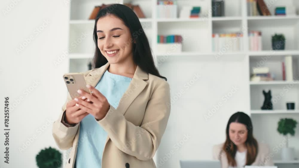Woman uses smart phone. Positive successful arabian or indian woman in a suit, stands in the office, holding mobile phone, texting in social media with friends or clients, making online order, smiles Woman uses smart phone. Positive successful arabian or indian woman in a suit, stands in the office, holding mobile phone, texting in social media with friends or clients, making online order, smiles