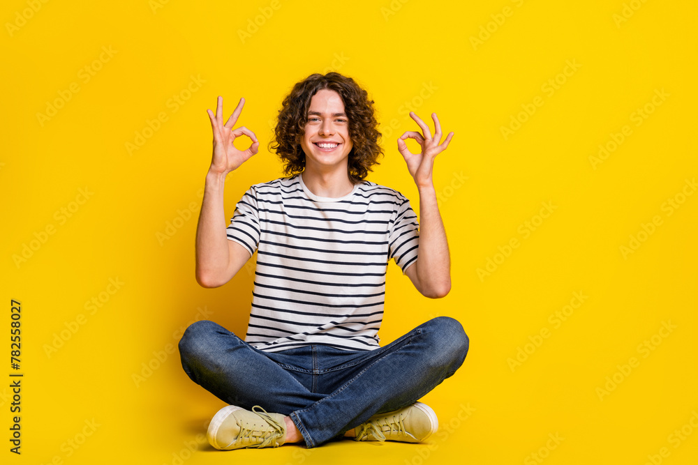 Full size portrait of nice young man sit floor show okey wear striped t-shirt isolated on yellow color background Full size portrait of nice young man sit floor show okey wear striped t-shirt isolated on yellow color background