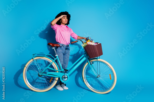 Full size photo of focused woman wear pink shirt hold bicycle look at promo empty space arm on forehead isolated on blue color background © deagreez