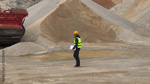 Quarry supervisor walking around the quarry in reflective vest and protective helmet with notebook in his arm. Walking past heavy machinery and large piles of gravel.