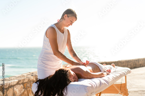 Close-up massage at beach on special table. Caucasian female masseur massages hand and back dark-haired tanned brunette woman. Tourist woman enjoying spa body treatment during vacation on ocean shore