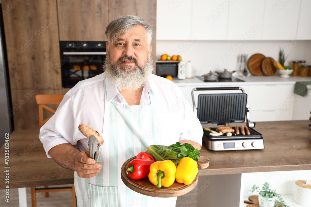 Mature man with board of fresh vegetables and tasty sausage cooked on modern electric grill at table in kitchen Mature man with board of fresh vegetables and tasty sausage cooked on modern electric grill at table in kitchen