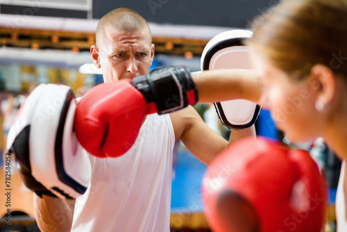 Young concentrated woman in boxing gloves launching blows on focus punch mitts during self defense workout in gym with trainer photo