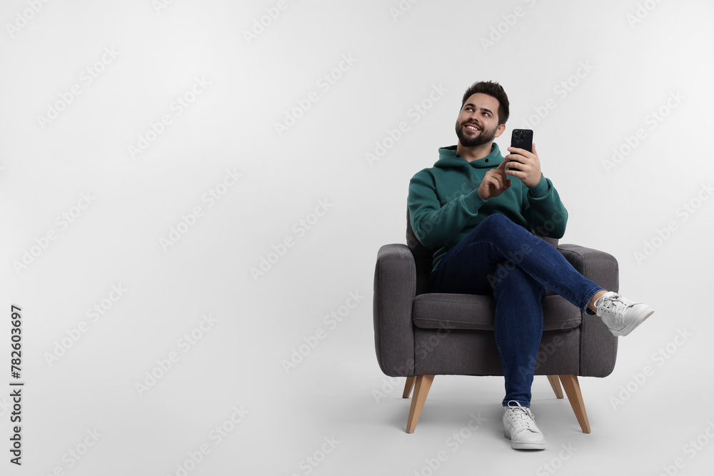 Happy young man using smartphone in armchair on white background, space for text