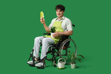 Young male gardener on wheelchair with houseplant and shovel on green background