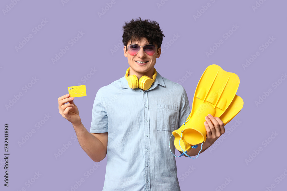 Young man in headphones holding credit card and flippers on lilac background Young man in headphones holding credit card and flippers on lilac background