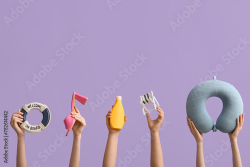 Female hands with travel pillow, sunscreen, miniature deck chair, heeled sandals and decorative lifebuoy on violet background. Travel concept.