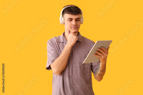 Handsome young thoughtful man in headphones with tablet listening audiobook on yellow background