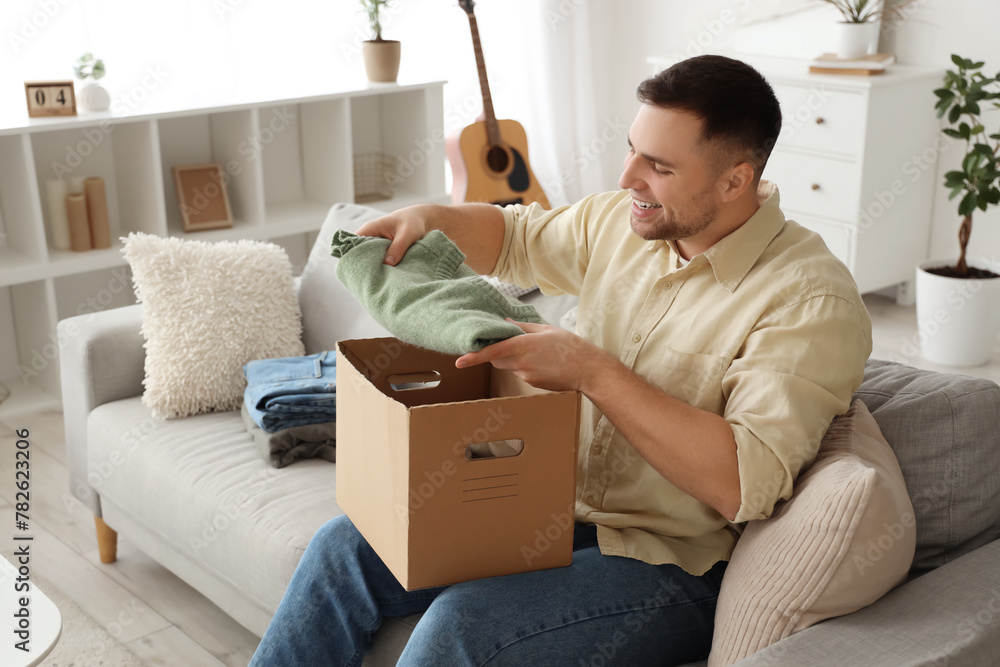 Young man packing folded clothes in box on sofa at home Young man packing folded clothes in box on sofa at home
