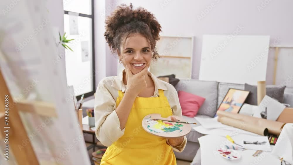 Thoughtful hispanic artist with curly hair, sitting, hand on chin in studio, lost in doubt, questioning expression on her face Thoughtful hispanic artist with curly hair, sitting, hand on chin in studio, lost in doubt, questioning expression on her face