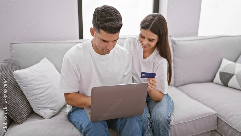 A smiling couple with a credit card and laptop comfortably shopping online together on a couch in a modern living room. A smiling couple with a credit card and laptop comfortably shopping online together on a couch in a modern living room.