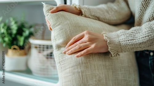 Close-up of person's hands gently holding beige textured cushion near window with light streaming in photo