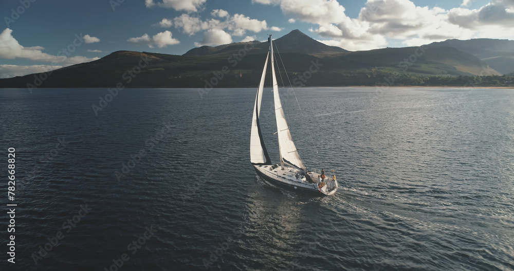 Aerial of sun yacht sail in ocean bay. White boat at open sea. Summer cruise on sailboat at sunlight with clouds. Scotland island of Arran sea coast with water transport. Serene seascape