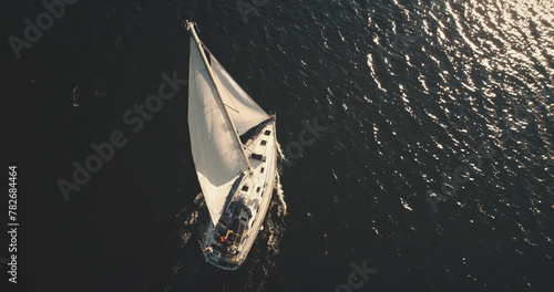 Top down of yacht under white sail at dark open sea aerial. Ocean bay black water and lonely sailboat. Summer cruise on boat. Luxury yachting scenery at serene seascape in dramatic drone shot
