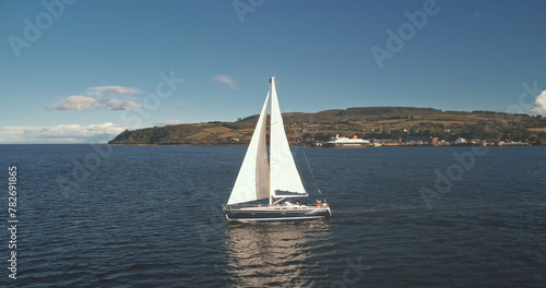 Sailboats cruise at open sea. Brodick port, Arran Island, Scotland. Nature seascape at summer day. Yachting at cargo terminal on ocean harbor aerial. Regatta on luxury yacht. Cinematic drone shot