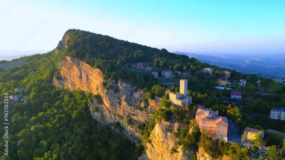Vivid aerial push-in shot from Montefalcone Appennino (Fermo) during the golden hour, with an impressive vegetation-filled mountainous cliff, roads, cars, buildings, and placid hills in the background Vivid aerial push-in shot from Montefalcone Appennino (Fermo) during the golden hour, with an impressive vegetation-filled mountainous cliff, roads, cars, buildings, and placid hills in the background