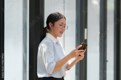 Happy Asian businesswoman in glasses standing smiling looking at data Talk on the phone using your smartphone for communication. Check through the application Go online while in the modern office.
