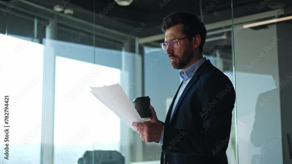 Side view of successful male in glasses reading report while drinking hot beverage indoors. Caucasian businessman learning information from documents and enjoying coffee in office environment. Side view of successful male in glasses reading report while drinking hot beverage indoors. Caucasian businessman learning information from documents and enjoying coffee in office environment.