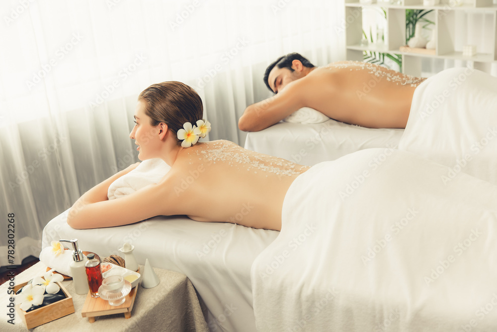 Blissful couple customer having exfoliation treatment in luxury spa salon with warmth candle light ambient. Salt scrub beauty treatment in health spa body scrub. Quiescent - obrazy, fototapety, plakaty 