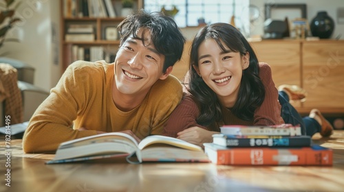 A Korean couple smiles happily with a stack of books while lying on their backs in the living room, looking at the ceiling. The man was lying on the left-hand side of the living room floor. The woman 