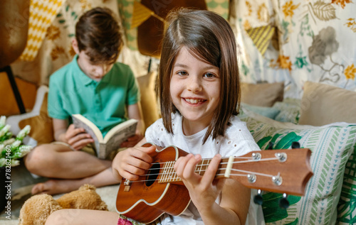 Portrait of smiling little girl looking at camera and playing ukulele while boy reading book on handmade teepee. Children having fun in diy shelter tent at home. Vacation camping or staycation concept