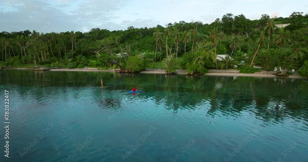 A Couple On Moso Island, Vanuatu. The Female Is On Paddleboarding While The Male Is On The Kayak Boat. Aerial Drone Shot A Couple On Moso Island, Vanuatu. The Female Is On Paddleboarding While The Male Is On The Kayak Boat. Aerial Drone Shot