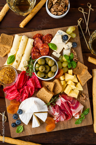 Charcuterie board with a variety of cheeses, salami, chorizzo, prosciutto, honey, grapes, nuts, olives, bread, blueberries and fresh herbs on a dark wooden background. A festive snack.