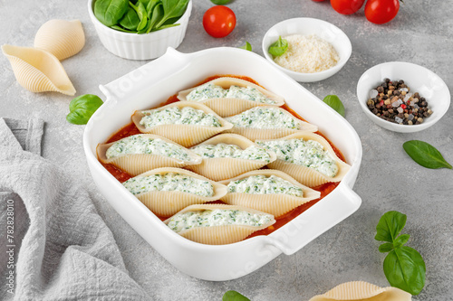 Raw conchiglioni pasta stuffed ricotta cheese and spinach with tomato sauce in a white baking dish on a gray concrete background.