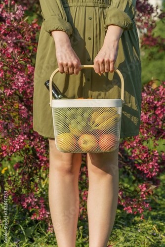 girl in simple green cotton dress with rolled-up sleeves holds white iron basket filled with fruits in close-up. concept of agriculture, environmental care, zero waste, reusable items, eco-friendly © Маргарита Трушина